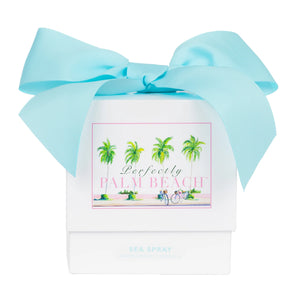 White box labeled "Perfectly Palm Beach" features palm trees, a bike, and a large blue bow. Inside is the "Sea Spray Candle," offering Palm Beach vibes with sea salt and hibiscus notes. Brand: Perfectly Palm Beach.