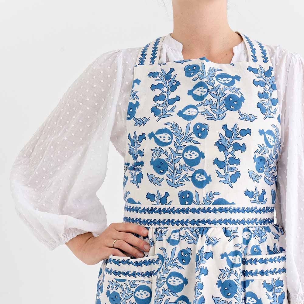 Wearing a white blouse with sheer polka dot sleeves, someone dons an adjustable apron from the Pomegranate Inc Apron Collection, featuring a blue floral design—ideal as a kitchen gift.