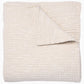 A folded cream-colored quilt from the Vivada Bedding Collection by John Robshaw features a hand-stitched, textured grid pattern with one corner flipped over to reveal its reversible design, beautifully capturing the timeless appeal of classic quilts.