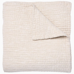 A folded cream-colored quilt from the Vivada Bedding Collection by John Robshaw features a hand-stitched, textured grid pattern with one corner flipped over to reveal its reversible design, beautifully capturing the timeless appeal of classic quilts.