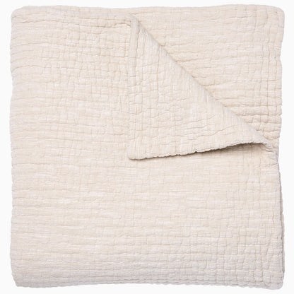 A folded cream-colored quilt from the Vivada Bedding Collection by John Robshaw features a hand-stitched, textured grid pattern with one corner flipped over to reveal its reversible design, beautifully capturing the timeless appeal of classic quilts.