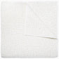 A white coverlet from the Hand-Stitched Bedding Collection by John Robshaw, featuring a subtle stitched pattern and organic cotton, is neatly folded with one corner turned down to reveal its detailed texture.