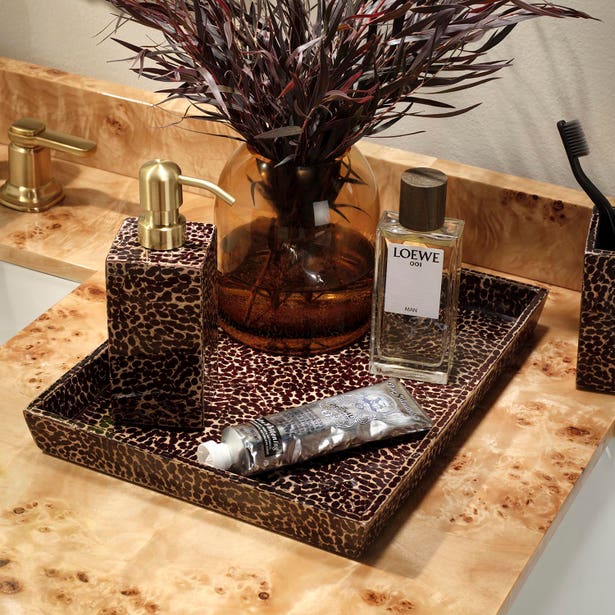 A bathroom counter features a leopard-print tray from the Callas Bath Collection by Pigeon & Poodle, neatly displaying a soap dispenser, hand cream, perfume, and a brown glass vase with foliage. A toothbrush appears in the background among these stylish essentials.