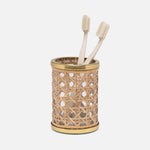 A cylindrical woven rattan toothbrush holder from the Norwell Bath Collection by Pigeon & Poodle, featuring gold accents, holds two beige toothbrushes against a plain white background.