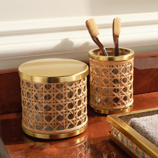 A cylindrical container with lid and a matching holder from the Pigeon & Poodle Norwell Bath Collection, featuring woven rattan and gold accents, sit on a wooden countertop holding two toothbrushes.