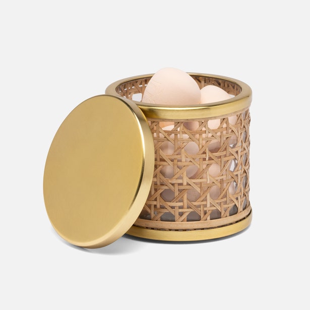 A round container from the Norwell Bath Collection by Pigeon & Poodle, crafted with woven rattan and a gold-colored rim and lid, sits partially open to reveal several eggs inside.