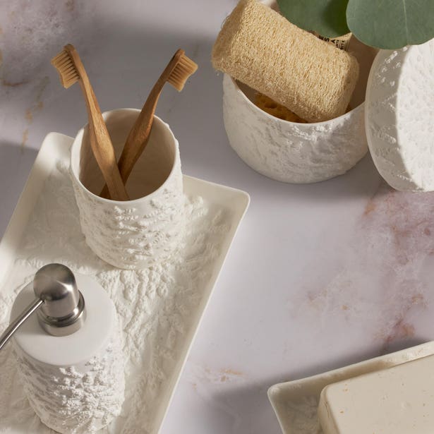 The Alanya White Bath Collection by Pigeon & Poodle features a coral-patterned porcelain soap dispenser, toothbrush holder with two wooden toothbrushes, soap dish and bar, loofah, and a vase with green leaves on marble.