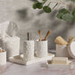 The Alanya White Bath Collection by Pigeon & Poodle showcases coral-patterned accessories—soap dispenser, toothbrush cups, tissue box, soap dish, and container—tastefully arranged on marble with leafy décor accents.