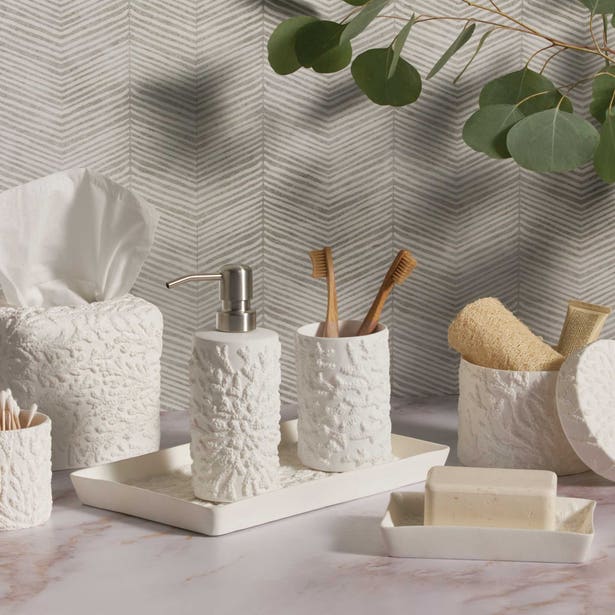 The Alanya White Bath Collection by Pigeon & Poodle showcases coral-patterned accessories—soap dispenser, toothbrush cups, tissue box, soap dish, and container—tastefully arranged on marble with leafy décor accents.