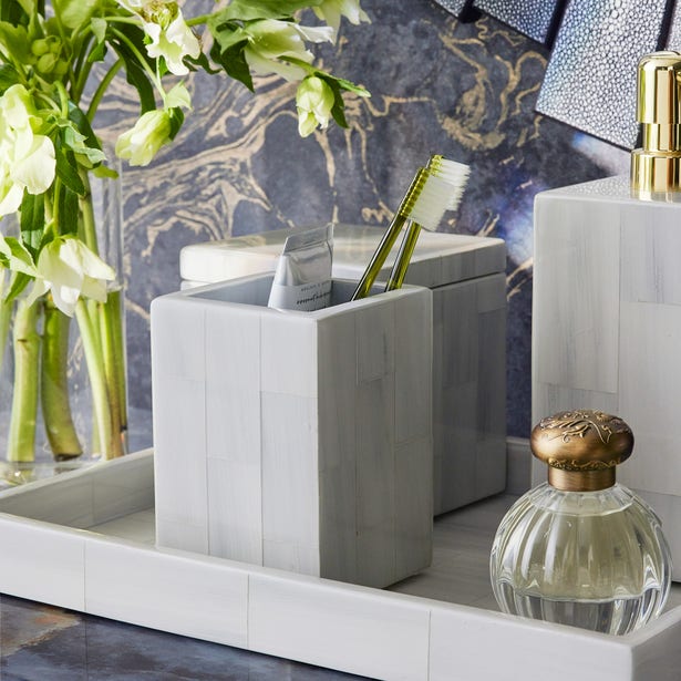The Arles Bath Collection by Pigeon & Poodle features white bathroom accessories, including a smooth toothbrush holder with two brushes, toothpaste, a glass jar with gold lid, and a vase of white flowers on a marble countertop.