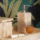 A stone soap dispenser, lidded container, glass perfume bottle, and rectangular box from Pigeon & Poodle's Bowen Natural Bath Collection sit on a stone tray against a leafy patterned background.