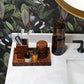A marble bathroom counter displays Pigeon & Poodle’s Carmel High Gloss Faux Tortoise accessories—including a toothbrush holder, tray, and bottles—set against botanical wallpaper for a distinct look.