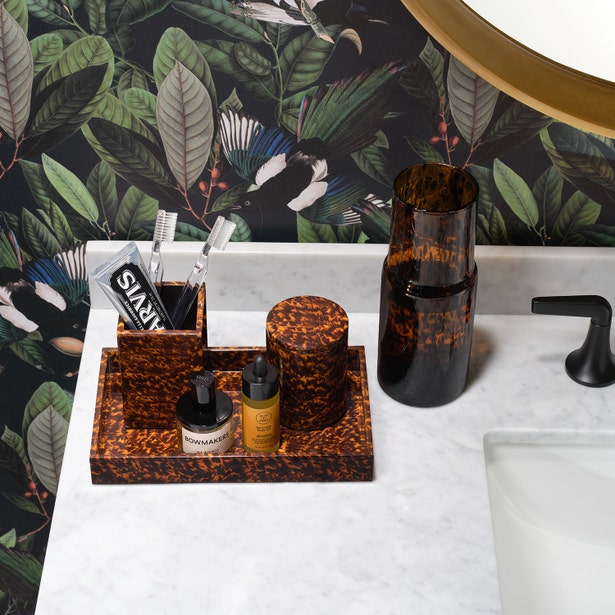 A marble bathroom counter displays Pigeon & Poodle’s Carmel High Gloss Faux Tortoise accessories—including a toothbrush holder, tray, and bottles—set against botanical wallpaper for a distinct look.