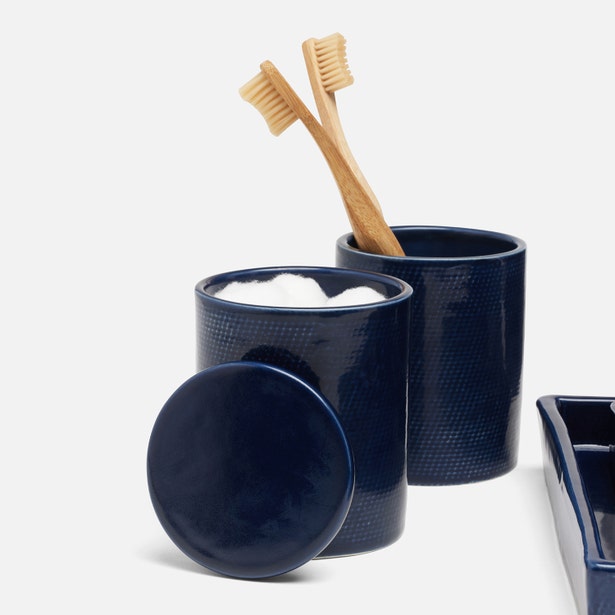 Two wooden toothbrushes sit in a navy ceramic cup from the Pigeon & Poodle Cordoba Navy Bath Collection, beside a hand-textured lidded cup filled with cotton balls, all arranged against a crisp white backdrop.