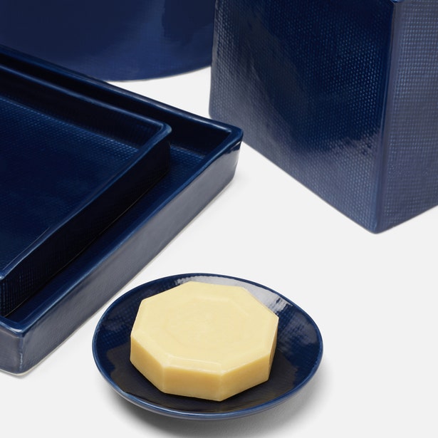 A hand-textured hexagonal bar of soap sits on a blue dish, with coordinating Cordoba Navy Bath Collection trays and containers by Pigeon & Poodle in the background.