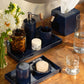 A bathroom countertop featuring Pigeon & Poodle’s Cordoba Navy Bath Collection ceramic toiletry set, with toothbrushes, soap, tissues, flowers in a vase, a glass of water, and a jar with a tortoiseshell lid.
