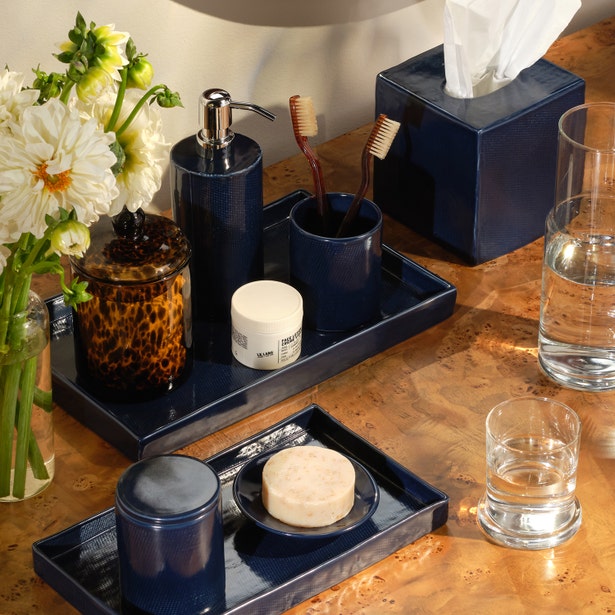 A bathroom countertop featuring Pigeon & Poodle’s Cordoba Navy Bath Collection ceramic toiletry set, with toothbrushes, soap, tissues, flowers in a vase, a glass of water, and a jar with a tortoiseshell lid.