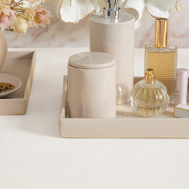 A beige countertop displays the Cordoba Bath Collection by Pigeon & Poodle—a matching tray with soap dispenser, jar, two perfume bottles, and tube—accented by dried flowers in a vase nearby.