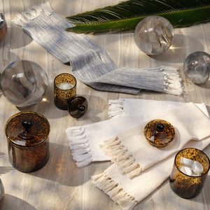 Two fringed cloth napkins, one gray and one white, are paired with handblown glass spheres, palm leaves, and the Pigeon & Poodle Tortoise Darby Canister on a light wooden surface.