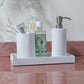 A white textured tray from the Dover Bath Collection by Pigeon & Poodle displays a cup with toothbrushes and toothpaste, a green bottle, and an engraved resin soap dispenser on a pink countertop.