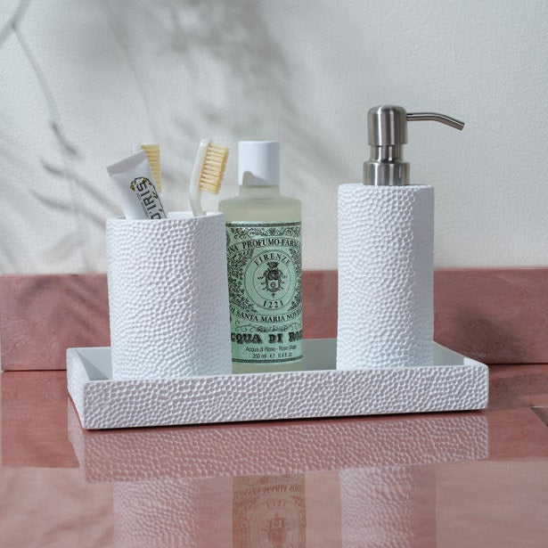 A white textured tray from the Dover Bath Collection by Pigeon & Poodle displays a cup with toothbrushes and toothpaste, a green bottle, and an engraved resin soap dispenser on a pink countertop.