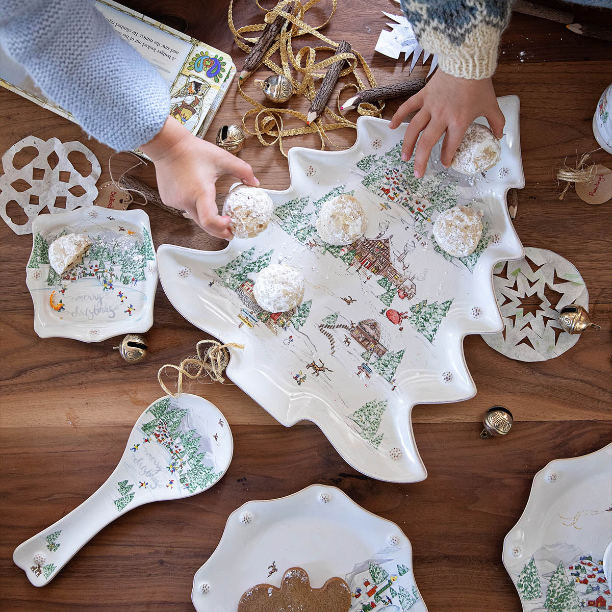 Two people arrange powdered cookies on a Juliska Berry & Thread North Pole Tree Platter Collection, surrounded by festive decor and ceramic dishes on a wooden table for a stunning winter wonderland display.