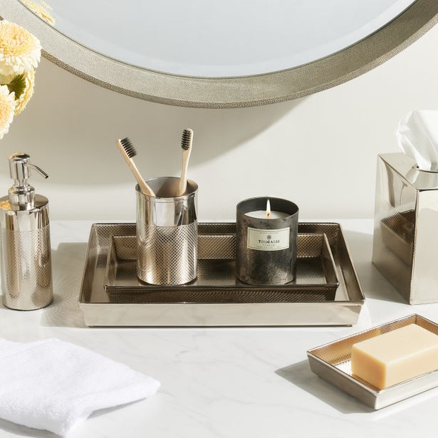 A shiny nickel tray from the Hagen Bath Collection by Pigeon & Poodle holds two toothbrushes, a metal cup, candle, and soap dispenser. A matching soap dish, tissue box, towel, and flower vase complete this elegant countertop display.