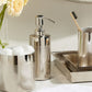 A bathroom countertop featuring the Hagen Bath Collection by Pigeon & Poodle: a stainless steel soap dispenser, a toothbrush cup, cotton ball container, tray, and a vase of light yellow flowers.