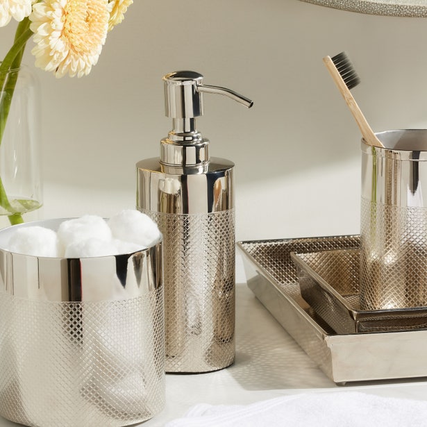 A bathroom countertop featuring the Hagen Bath Collection by Pigeon & Poodle: a stainless steel soap dispenser, a toothbrush cup, cotton ball container, tray, and a vase of light yellow flowers.