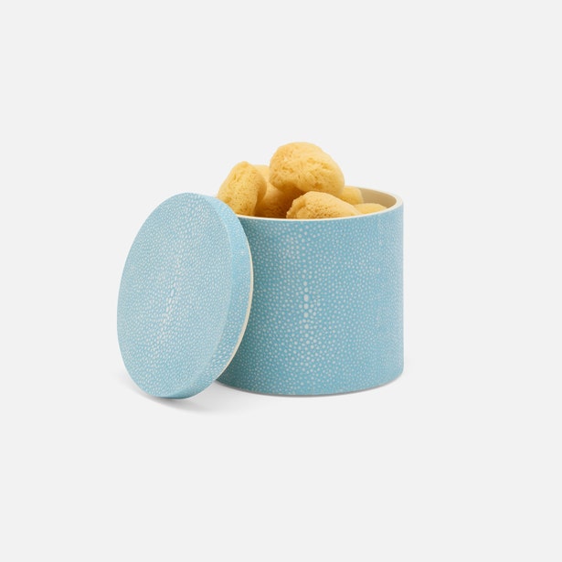 The Manchester Bath Collection by Pigeon & Poodle features a light blue cylindrical container with a matching lid, subtly decorated with a natural eye pattern and partially open to reveal round yellow cookies inside.