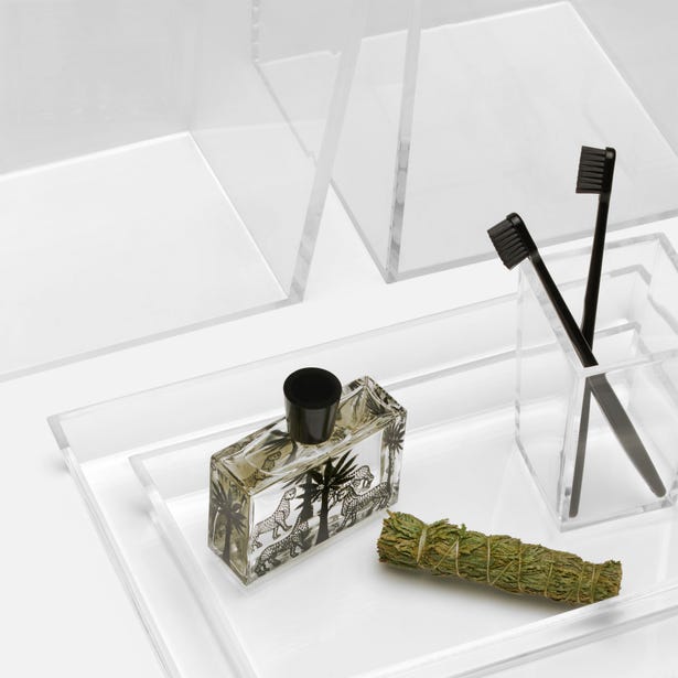 A clear tray from the Monette Bath Collection by Pigeon & Poodle holds a rectangular perfume bottle with a black cap, a bundle of sage, and a clear holder containing two black toothbrushes.