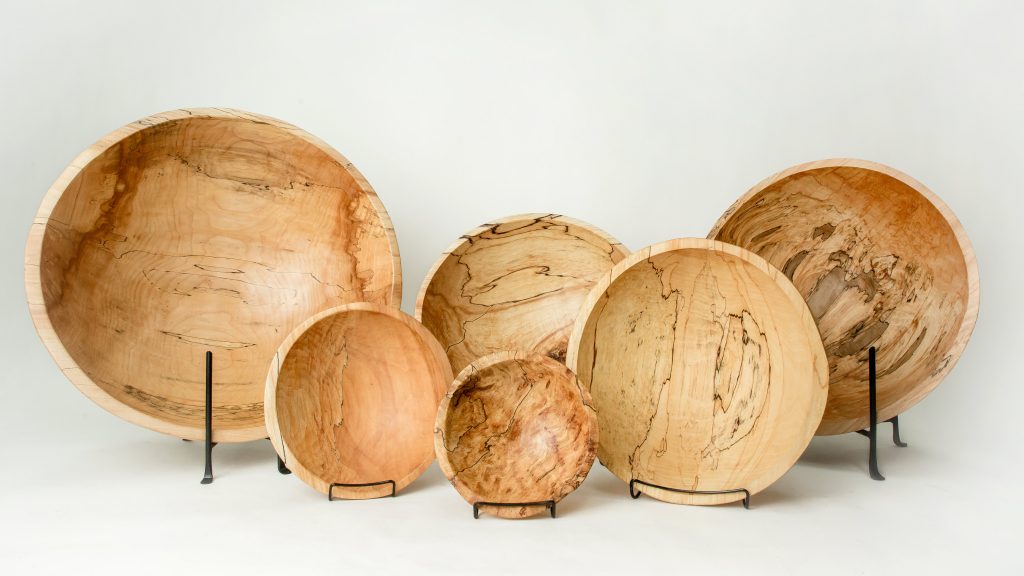 Peterman's Spalted Maple Serving Bowl Collection showcases six handcrafted wooden bowls of different sizes, each with distinct grain patterns, displayed upright on black stands against a white background.