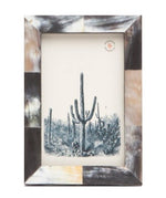 The Pigeon & Poodle Dark Horn Essen Frame, 4 x 6, features a checkered pattern in brown, black, and cream tones reminiscent of African horn and displays a black-and-white desert scene with large cacti.