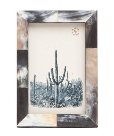 The Pigeon & Poodle Dark Horn Essen Frame, 4 x 6, features a checkered pattern in brown, black, and cream tones reminiscent of African horn and displays a black-and-white desert scene with large cacti.