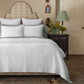 A neatly made bed features the John Robshaw Hand-Stitched Bedding Collection in white, complete with matching pillows, a patterned headboard, and is accompanied by a dark nightstand topped with a green lamp and a vase of yellow flowers.