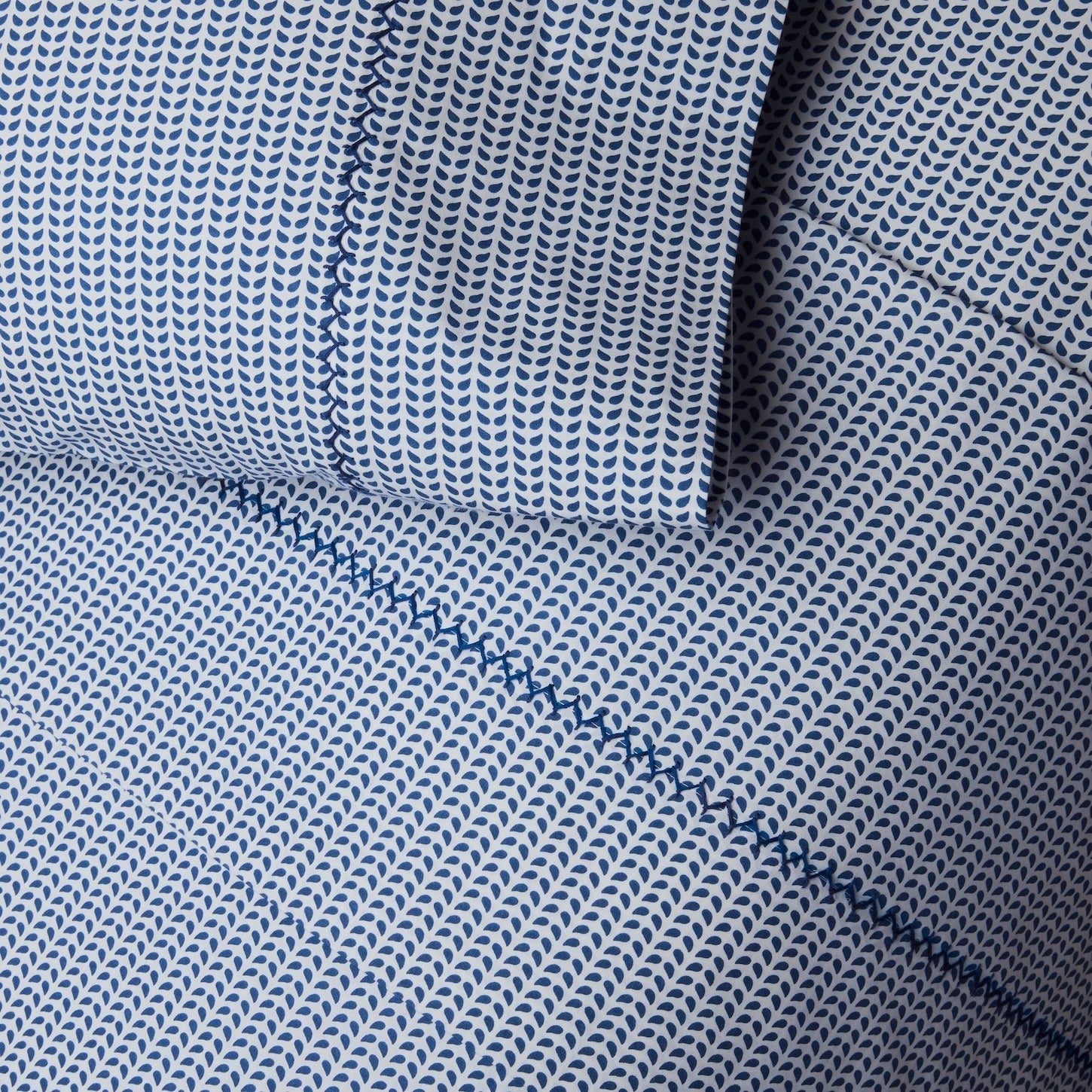 Close-up of a Light Indigo and white patterned organic sheet set from the Cinde Bedding Collection by John Robshaw, featuring small repeating leaf-like shapes in the signature Cinde print, with visible stitching detail.