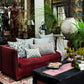 A red sofa adorned with the Mahisha Decorative Pillow by John Robshaw sits in a lush, eclectic living room filled with plants, artwork, books, and a large white vase near a window.