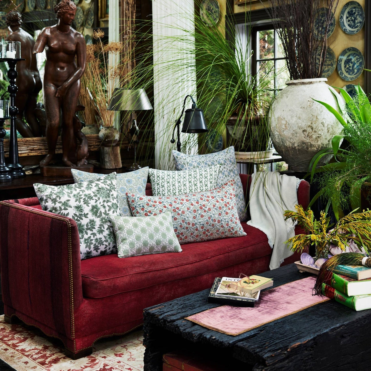 A red sofa adorned with the Mahisha Decorative Pillow by John Robshaw sits in a lush, eclectic living room filled with plants, artwork, books, and a large white vase near a window.