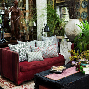 A red sofa adorned with the Mahisha Decorative Pillow by John Robshaw sits in a lush, eclectic living room filled with plants, artwork, books, and a large white vase near a window.