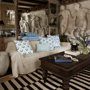 Living room with a white sofa adorned with cotton linen, accented by John Robshaw’s Ankur Azure Pillow—patterned in blue and white with delicate embroidered flowers and a hidden zipper closure—a dark wood coffee table, striped rug, flowers in a vase, and wall art featuring classical sculptures and framed photos.