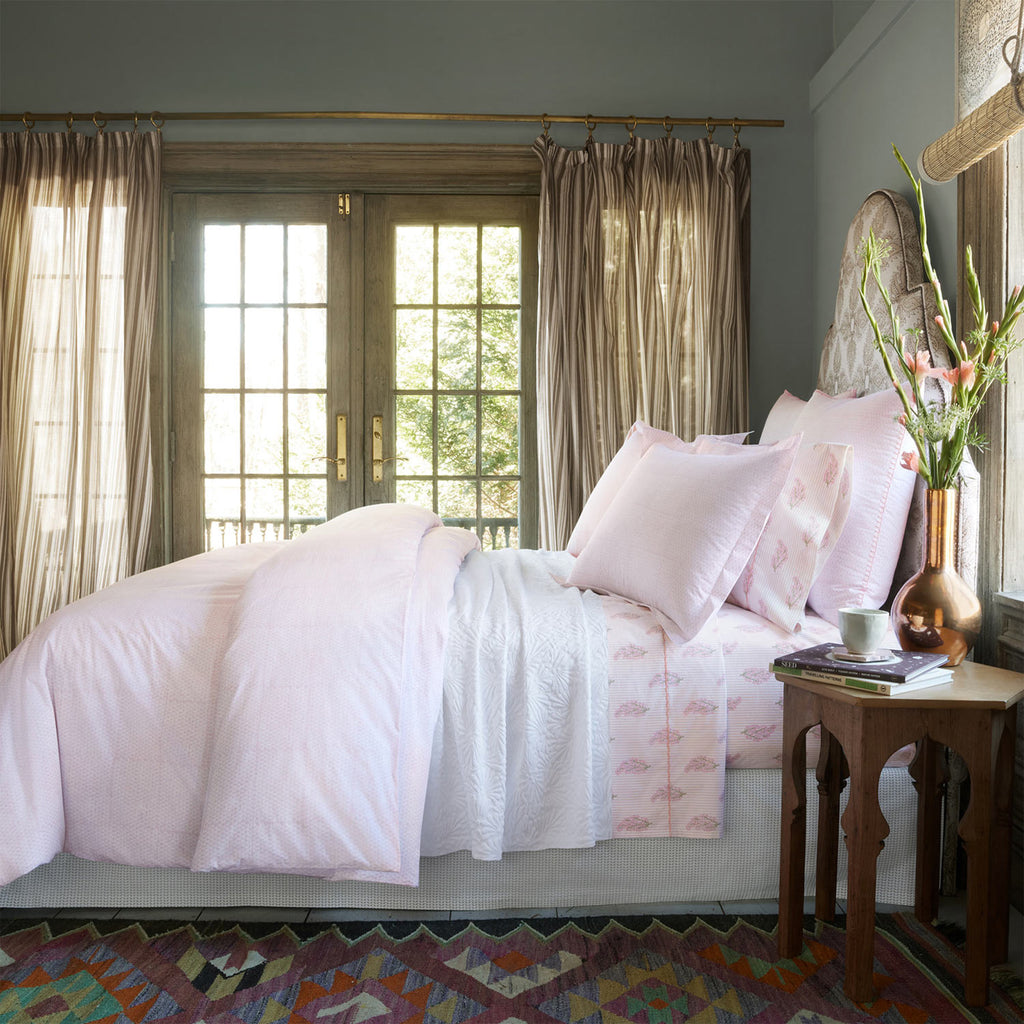 A neatly made bed with the Nirvi Lotus Bedding Collection by John Robshaw, showcasing pale pink subtle stripes, sits beside a small wooden nightstand with a vase of flowers in a sunlit room with sheer curtains and double glass doors.