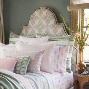 A neatly made bed with John Robshaw's Rohan Lotus Kidney Pillow, an embroidered upholstered headboard, and a nightstand with a vase of lotus blossoms by the window.