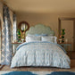 A neatly made bed with the John Robshaw Vidya Leaf Bedding Collection in blue and sage, flanked by side tables with vases of regal blooms, in a bright bedroom featuring patterned curtains and a decorative headboard.