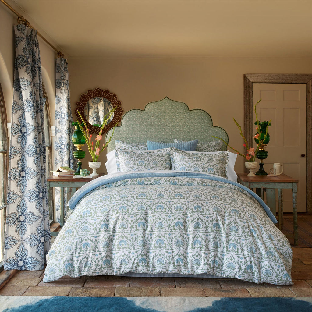 A neatly made bed with the John Robshaw Vidya Leaf Bedding Collection in blue and sage, flanked by side tables with vases of regal blooms, in a bright bedroom featuring patterned curtains and a decorative headboard.
