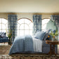 A bedroom featuring the Sahil Indigo Bedding Collection by John Robshaw, paired with matching curtains, a floral armchair, and a round wooden side table with a vase, all framed by arched windows.