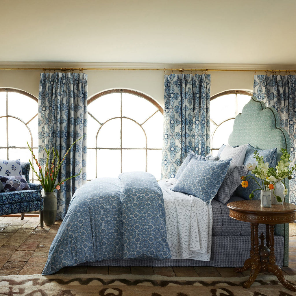 A bedroom featuring the Sahil Indigo Bedding Collection by John Robshaw, paired with matching curtains, a floral armchair, and a round wooden side table with a vase, all framed by arched windows.