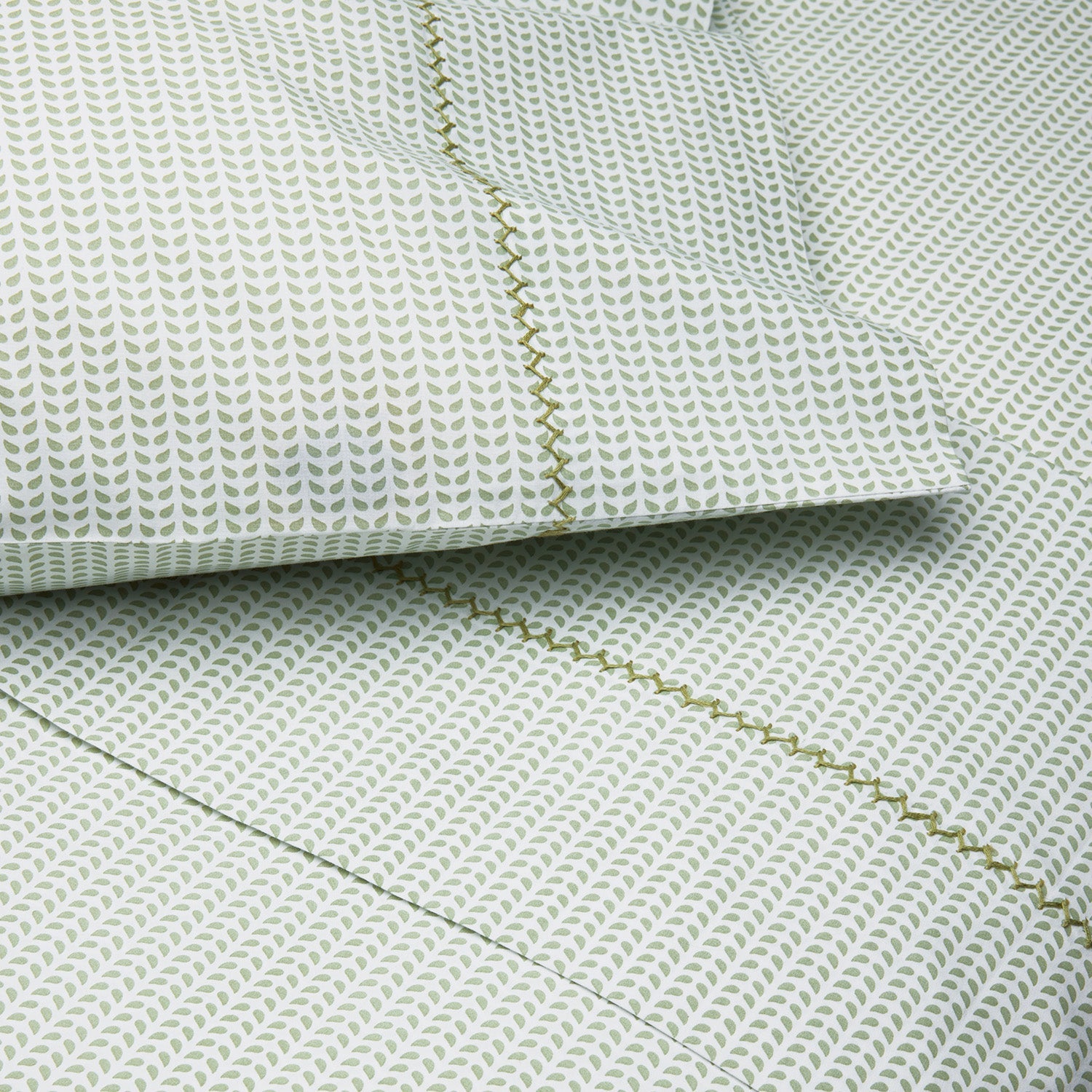 Close-up of a bed featuring the John Robshaw Cinde Bedding Collection, with green and white patterned sheets and pillowcases adorned with a small leaf motif and a decorative green stitched border. This organic sheet set also highlights the delicate Cinde print details for an added touch of style.