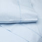 Folded and layered to reveal their textured fabric and signature Cinde print, these organic bed sheets from the John Robshaw Cinde Bedding Collection showcase a Light Indigo hue with small white leaf motifs and visible stitched seams.
