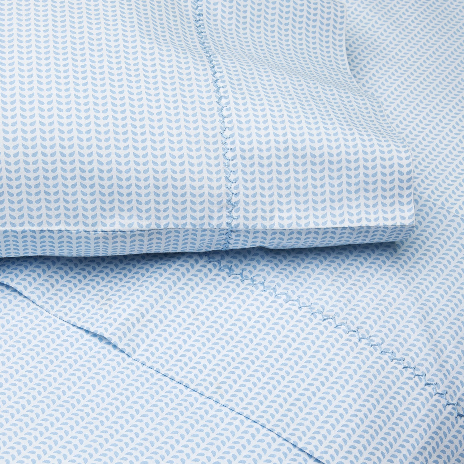 Folded and layered to reveal their textured fabric and signature Cinde print, these organic bed sheets from the John Robshaw Cinde Bedding Collection showcase a Light Indigo hue with small white leaf motifs and visible stitched seams.