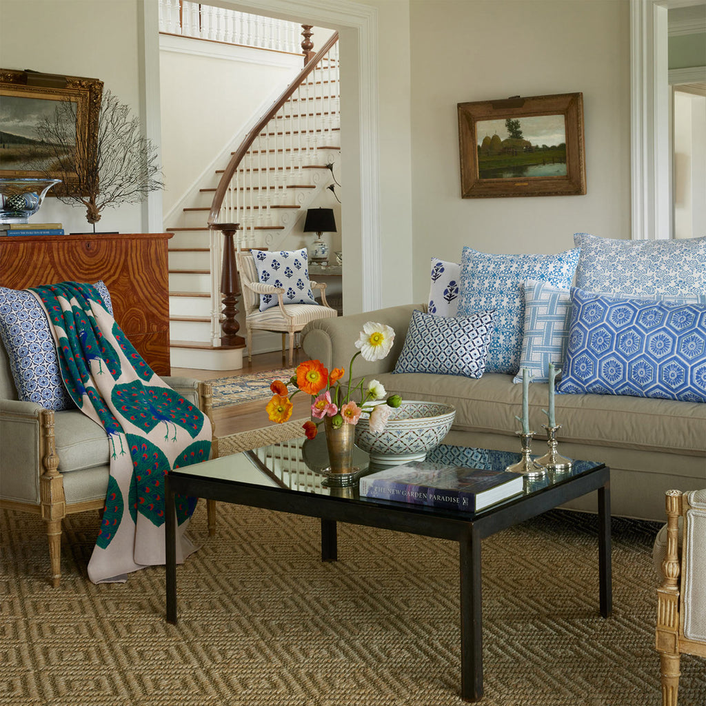 A living room features two beige sofas with John Robshaw’s Kripa Light Indigo Kidney Pillows, showcasing sky blue and indigo floral motifs. A glass coffee table holds books and flowers, with a staircase behind. French knot embroidery adds charming texture.